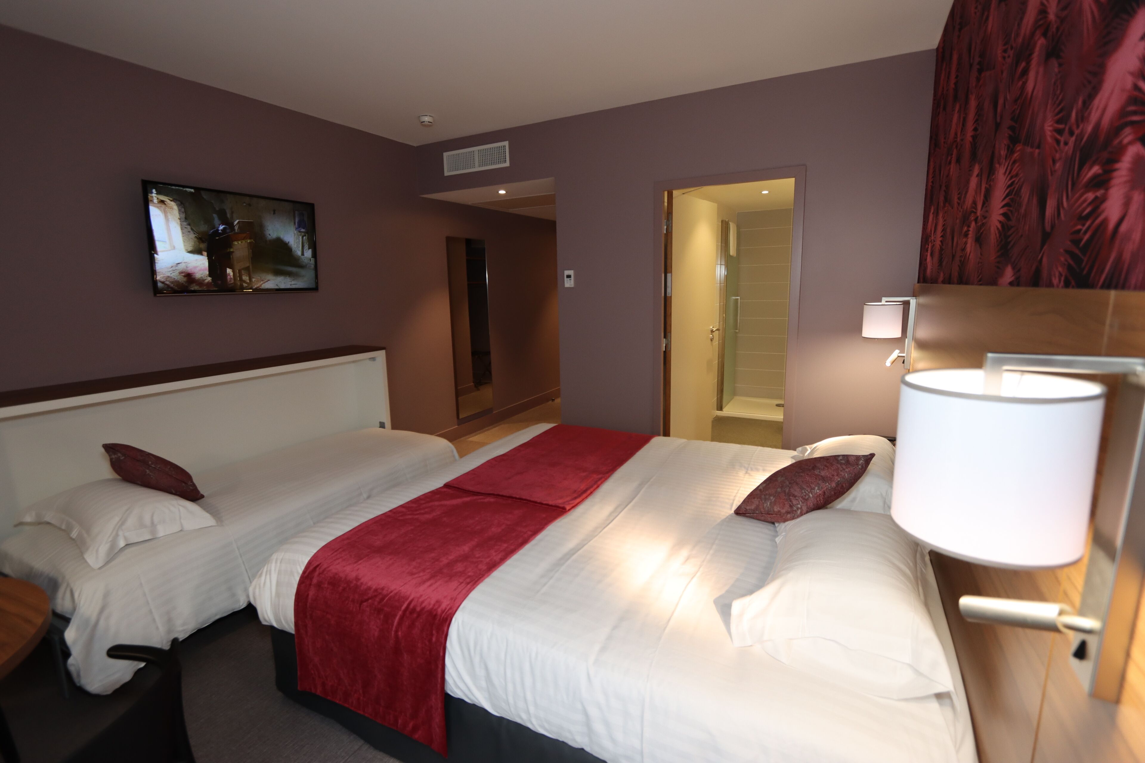 A hotel room with two beds, a TV on the wall, a red throw blanket, and a lamp.