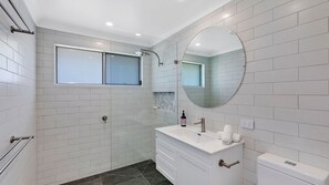 Un baño moderno con baldosas blancas, un espejo redondo, cabina de ducha de cristal y tocador blanco con lavamanos y toalla.