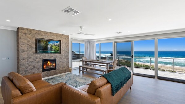 A modern living room with a fireplace, leather sofas, a dining area, and a large sliding glass door leading to a balcony with a view of the ocean.