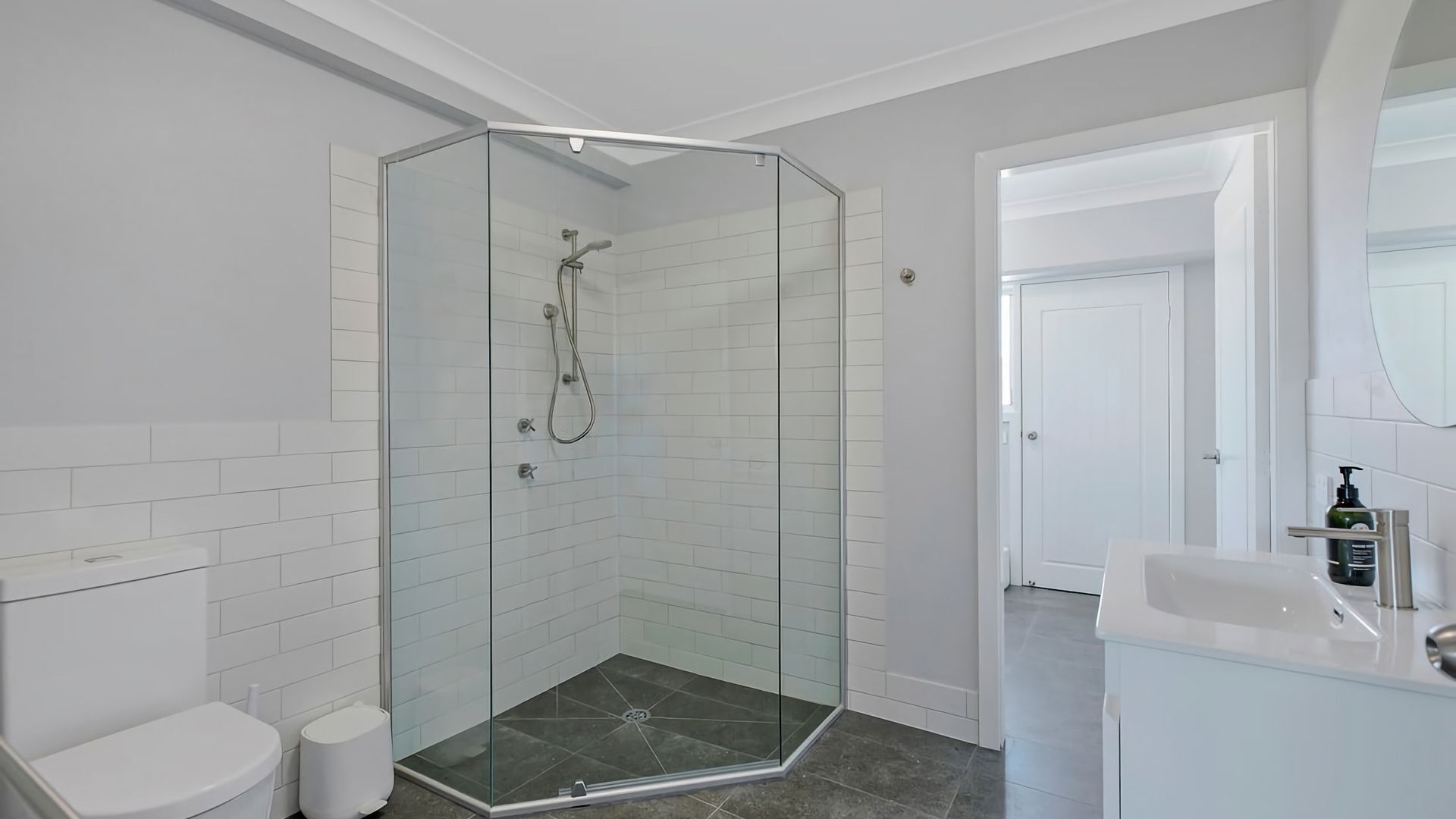 Une salle de bain moderne avec une douche à parois de verre, des carreaux blancs, des toilettes blanches et un lavabo avec un miroir.