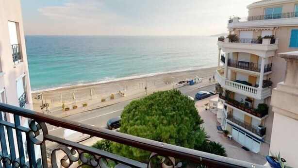 A coastal view from a balcony overlooking a beach, with a road in the foreground and a multi-story building with balconies. - Spacious 1-Bedroom Apartment with Sea View, Balcony, Pool, and Parking in Central Menton (Menton)