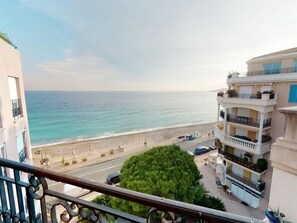 Uitzicht op de kust vanaf een balkon, met daaronder een strand, een weg op de voorgrond en een meerlaags gebouw met balkons.