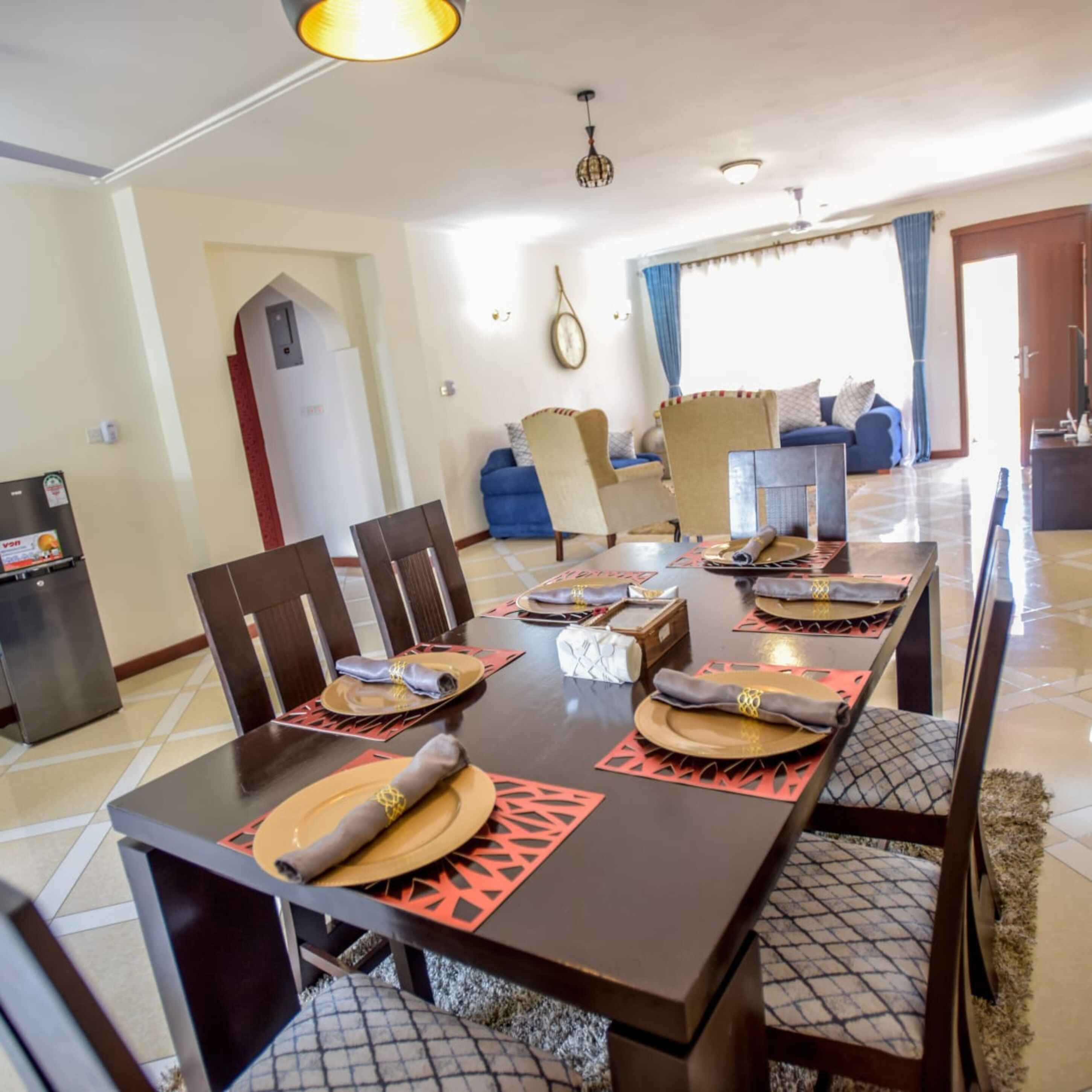 A dining area with a long table set for four, surrounded by chairs. There is a refrigerator and a door leading to another room.