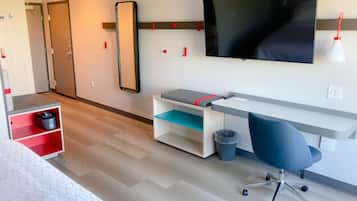 Une chambre d’hôtel moderne dotée d’un grand téléviseur à écran plat fixé au mur, d’un bureau avec une chaise, d’une petite poubelle et d’un meuble de rangement avec des étagères rouges.