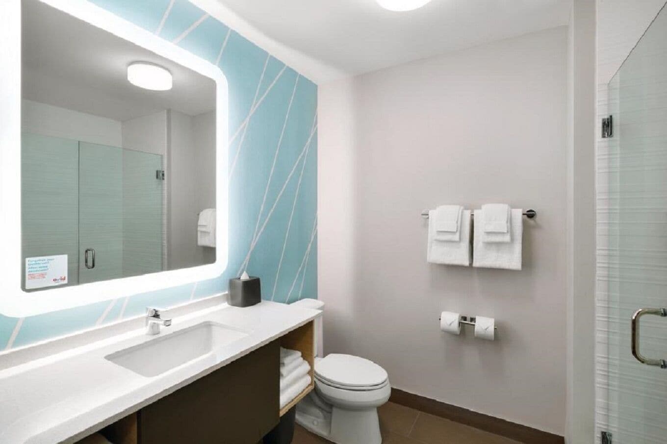 A modern bathroom with a large mirror, white sink, and a toilet. There are towels hanging on the wall and a glass shower enclosure.
