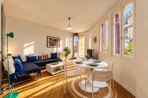 A living room with a sofa, a dining table set for two, and a large window with a view of buildings.