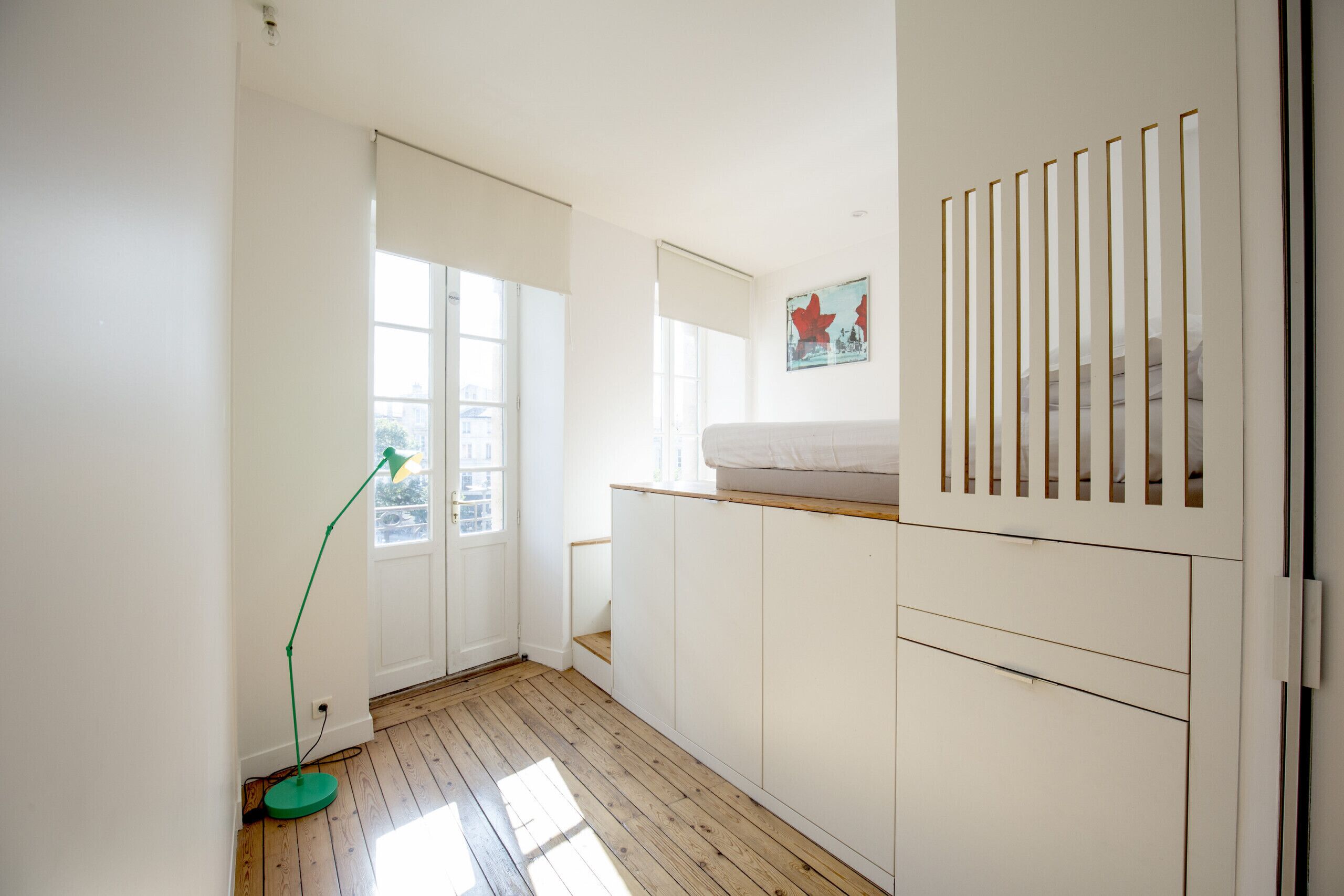 Een kamer met een houten vloer, witte muren, een groene vloerlamp, een bed met een witte matras en een raam met uitzicht op gebouwen.