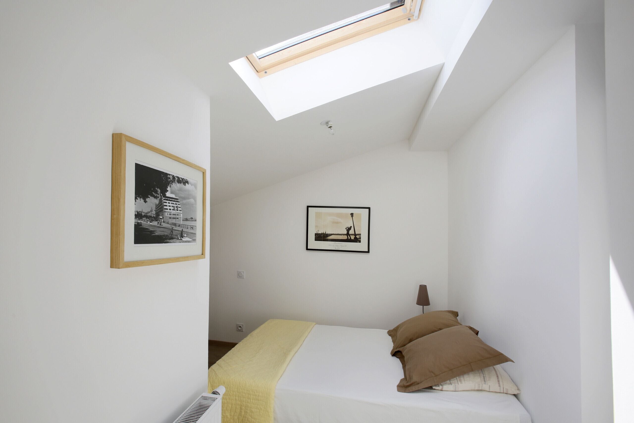 Un dormitorio pequeño y luminoso con tragaluz, una cama con almohadas y dos cuadros en la pared.