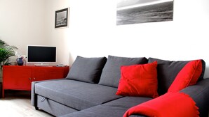 A modern living room with a grey sofa, red cushions, a flat-screen TV on a red cabinet, and a framed picture on the wall.