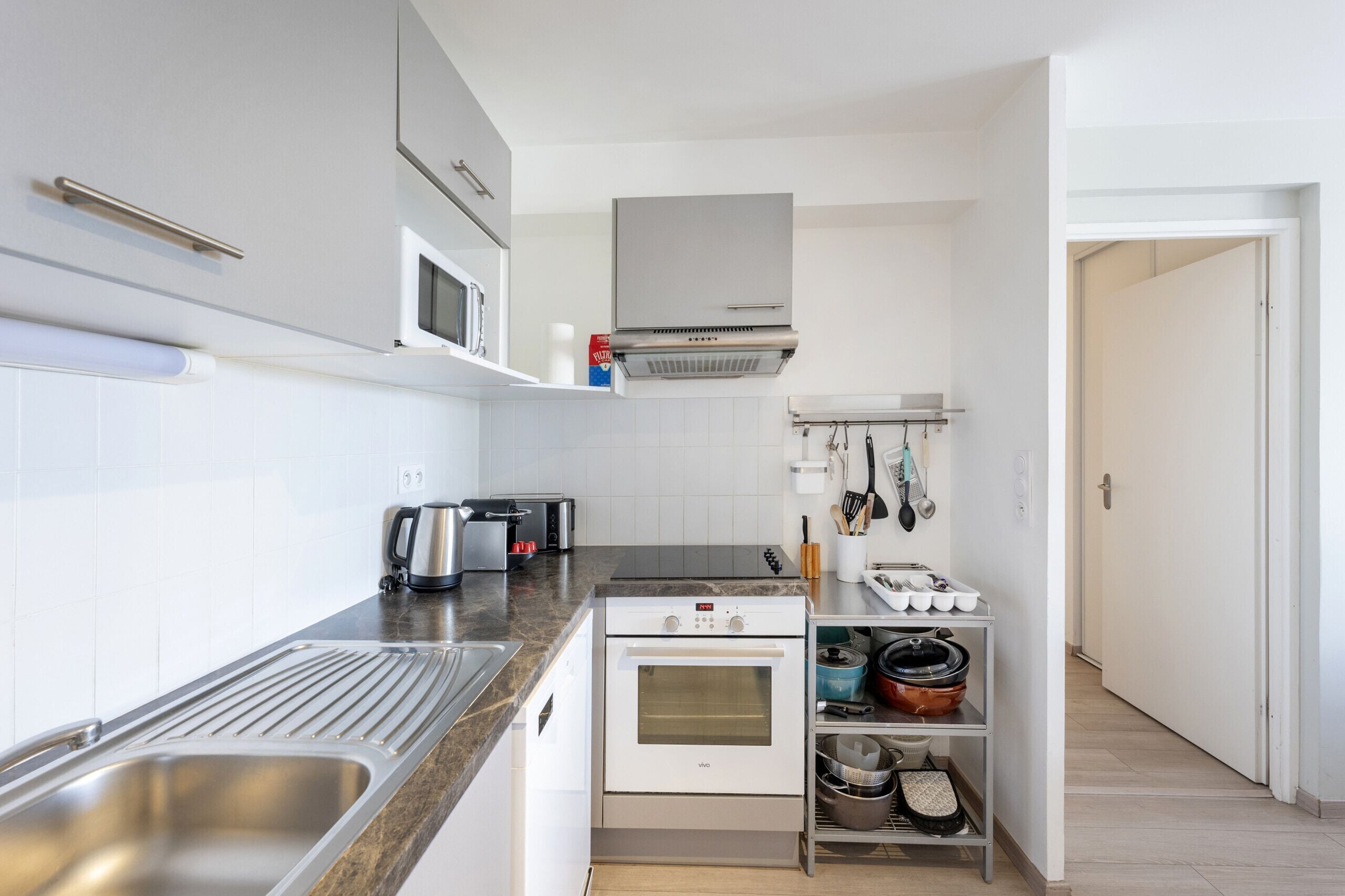 A compact kitchen with a stainless steel sink, a built-in oven, a microwave, and a range hood. There are cabinets above the counter and a shelf with cookware.