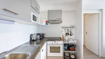 A compact kitchen with a stainless steel sink, a built-in oven, a microwave, and a range hood. There are cabinets above the counter and a shelf with cookware.