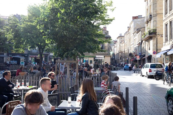 Un café en plein air, avec des tables et des chaises, entouré d’immeubles et d’un arbre.