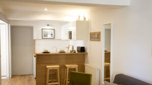 Een compacte leefruimte met een kitchenette, twee houten krukken, een groene stoel en een grijze bank met gele kussens.