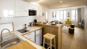Een moderne keuken met een spoelbak, fornuis, magnetron en een kleine koelkast. Er staat een houten kruk en er is een eethoek met een tafel en stoelen.