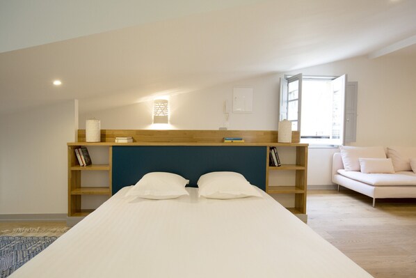 Ein Schlafzimmer mit einem Bett, einem hölzernen Kopfteil, einem Nachttisch mit Büchern, einem weißen Sofa und einem Fenster mit Tageslicht.