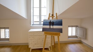 A room with a wooden floor, a white dresser, a painting of a couple on a beach, and a window with white curtains. - Bordeaux Historic Center Superior double studio (Burdeos)