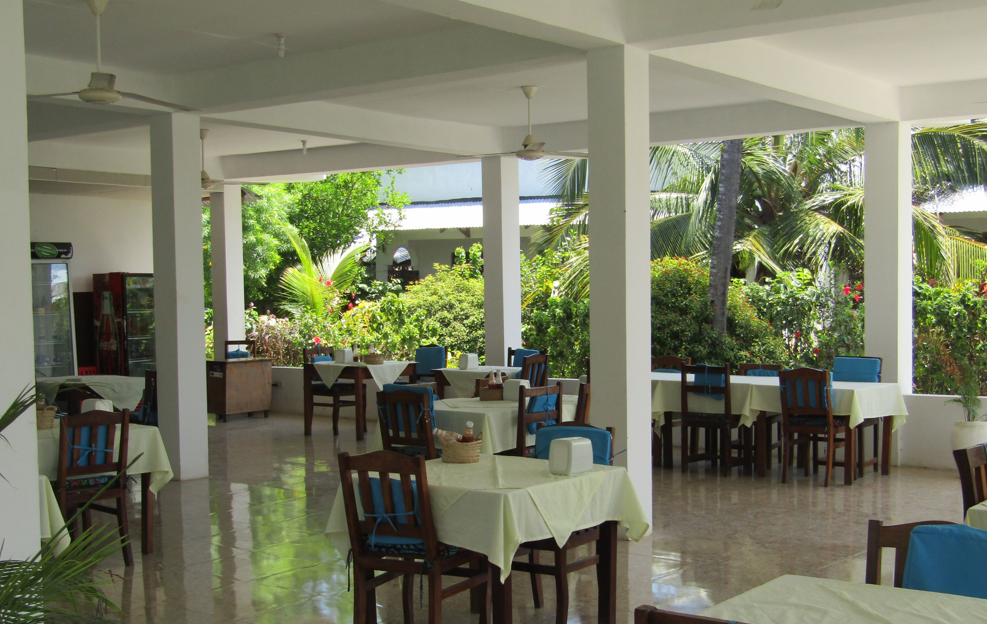Un vaste espace de restauration intérieur, doté de nombreuses tables et chaises, de grandes fenêtres offrant une vue sur une végétation luxuriante et de ventilateurs de plafond.