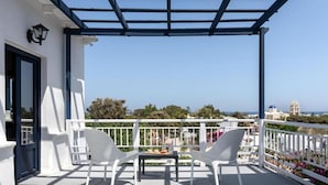 A balcony with two white wicker chairs and a small table, overlooking a scenic view of a coastal town with buildings and a clear sky. - Rivari Apartments (Santorini)