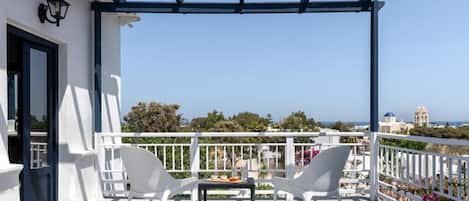A balcony with two white wicker chairs and a small table, overlooking a scenic view of a coastal town with buildings and a clear sky.