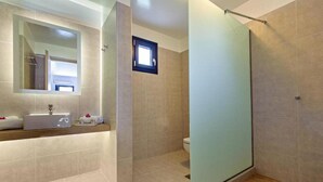 A modern bathroom with a glass shower enclosure, a large mirror, and a vanity with a sink and towel rack. - Rivari Apartments (Santorini)