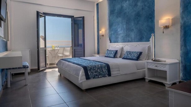 A modern bedroom with a large bed, a bedside table, a nightstand, and a view through an open door to a balcony. - Rivari Apartments (Santorini)