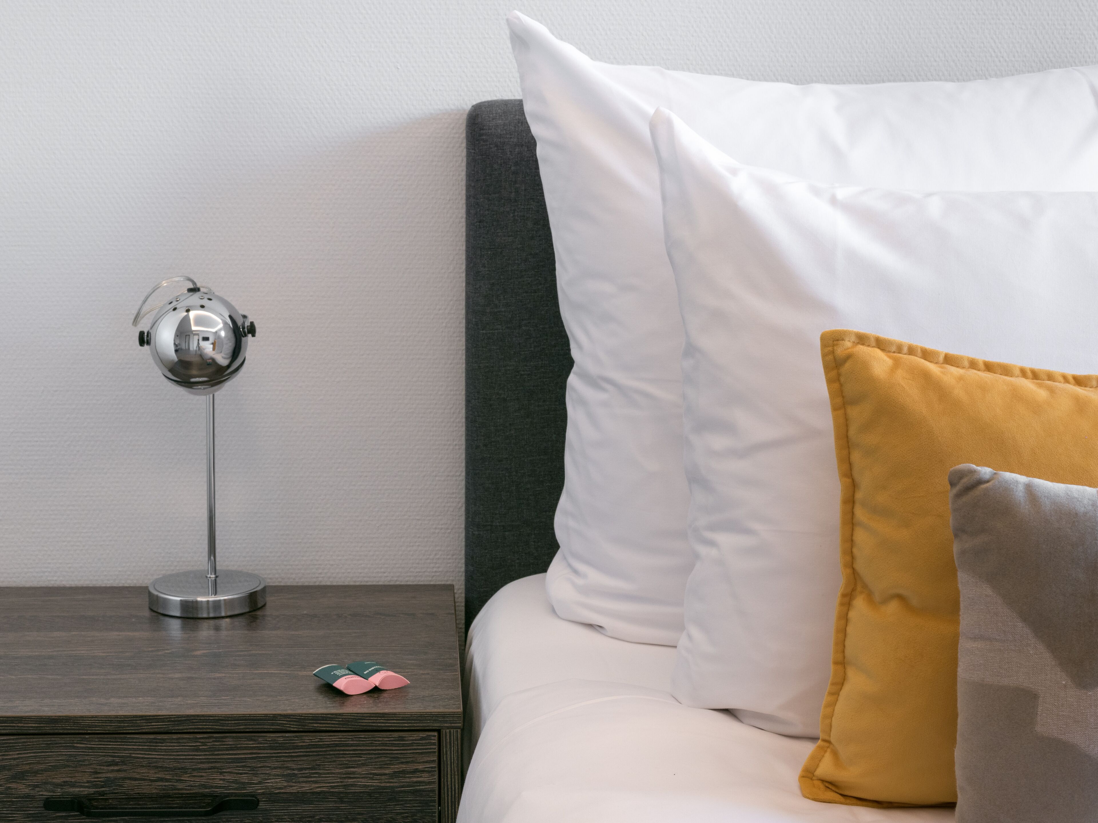 Una cama bien tendida con almohadas blancas y mostaza, una mesita de noche con una lámpara y un pequeño objeto decorativo.
