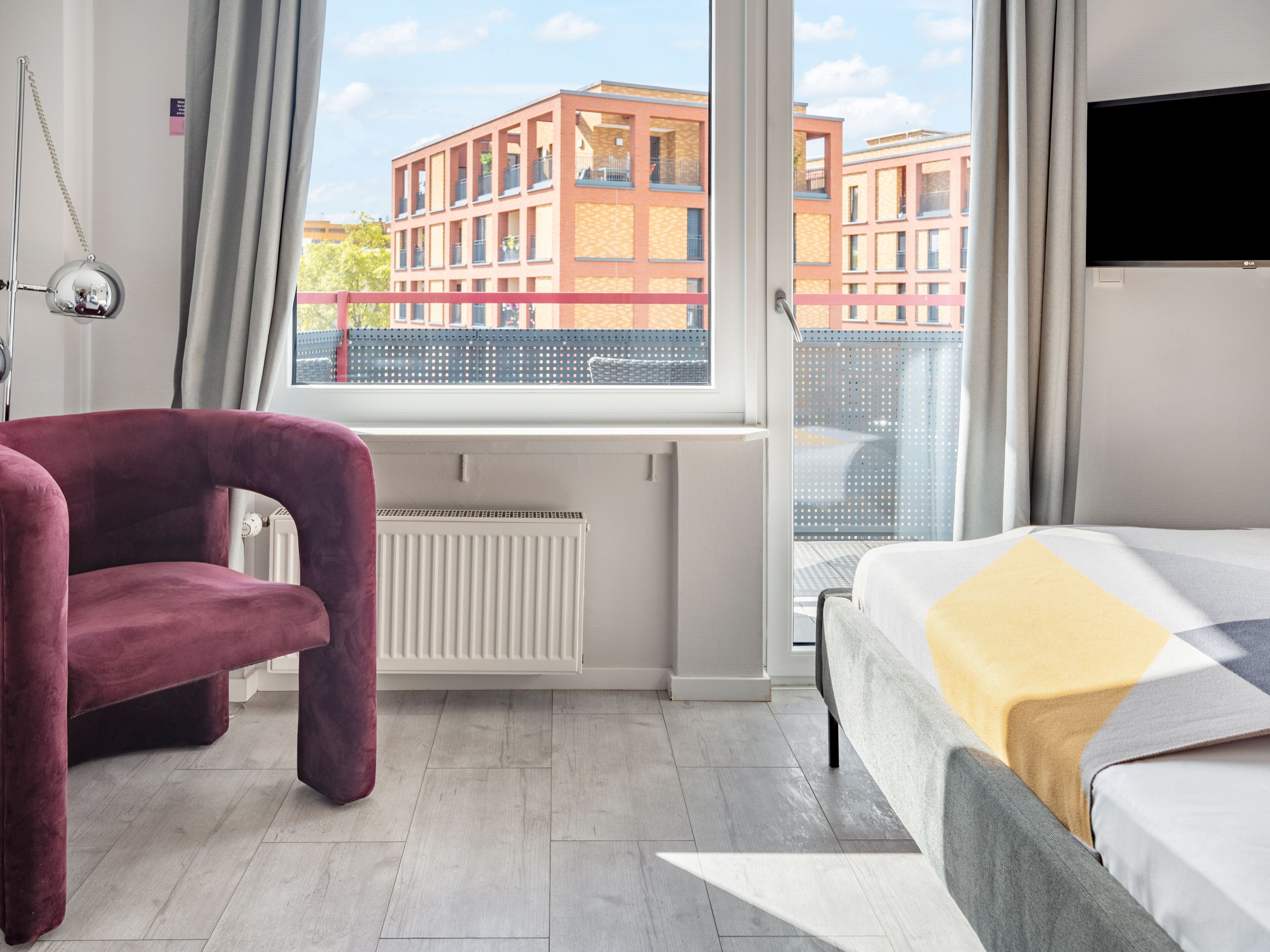 A modern hotel room with a large window, a red chair, a bed with a patterned bedspread, and a flat-screen TV.