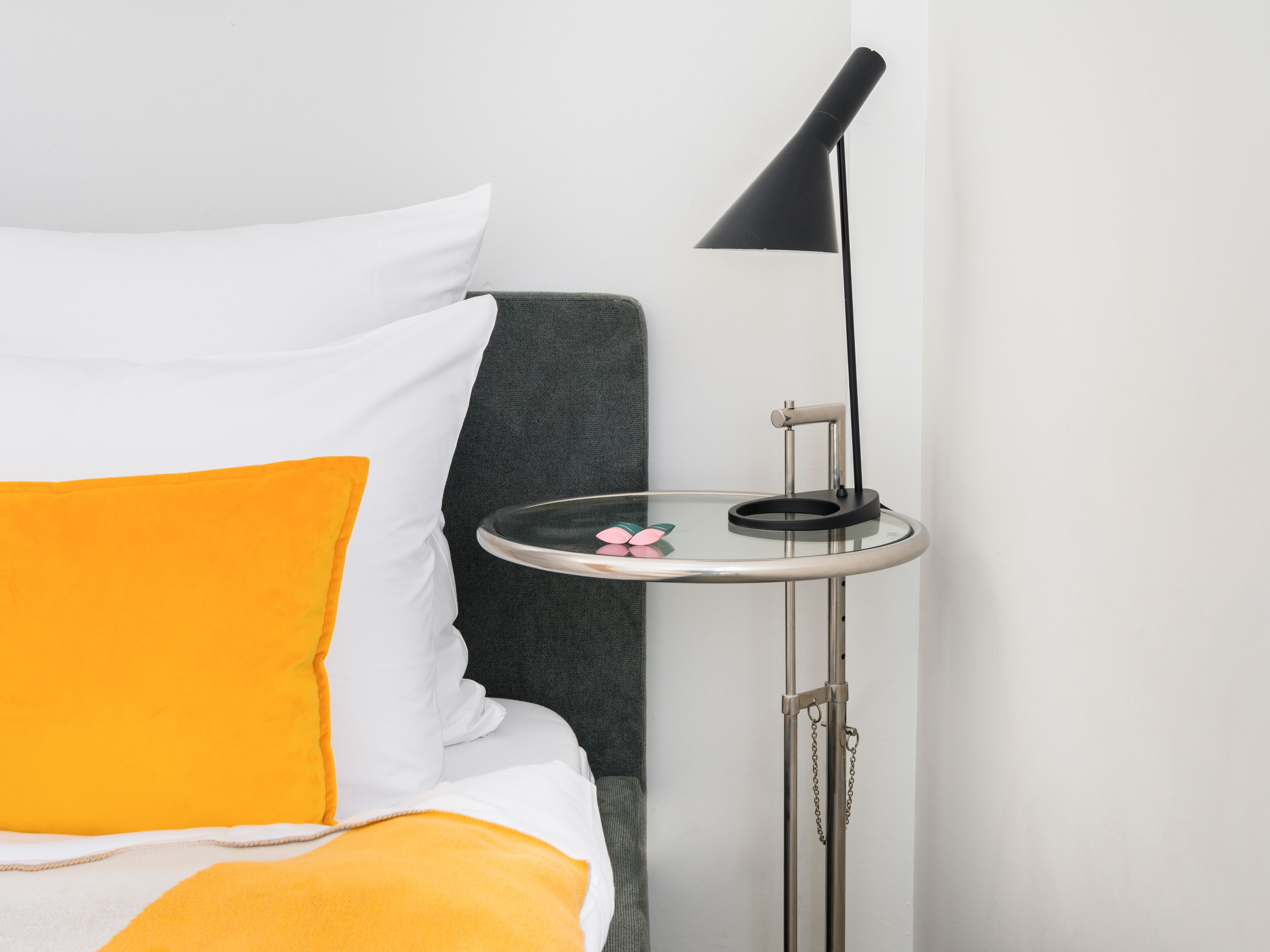 Uma cama bem arrumada com roupa de cama branca e laranja, uma cabeceira cinza e uma pequena mesa com um abajur preto ao lado.