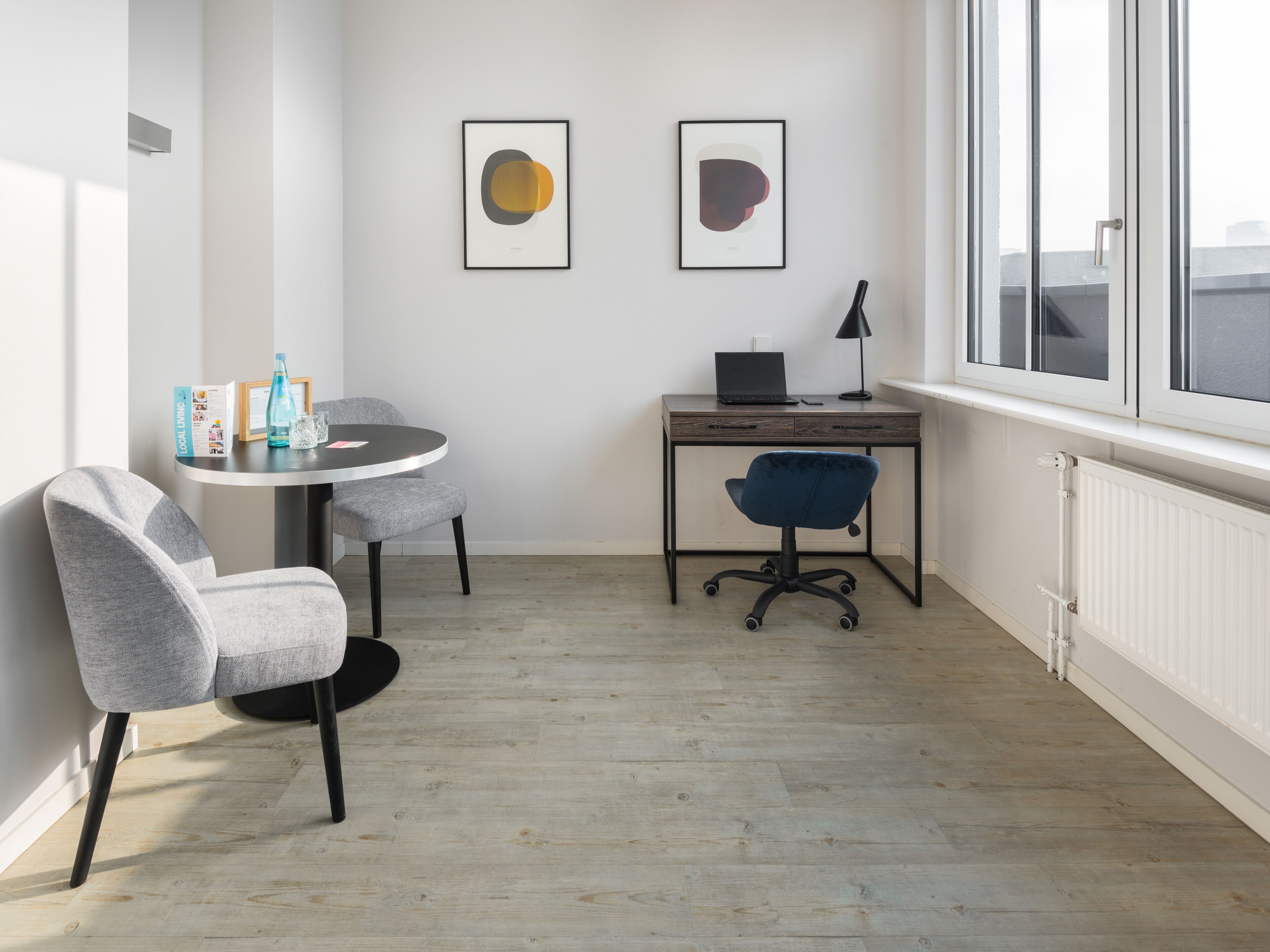 Een moderne kamer met een ronde tafel, twee stoelen, een bureau met een laptop en twee kunstwerken in lijsten aan de muur.