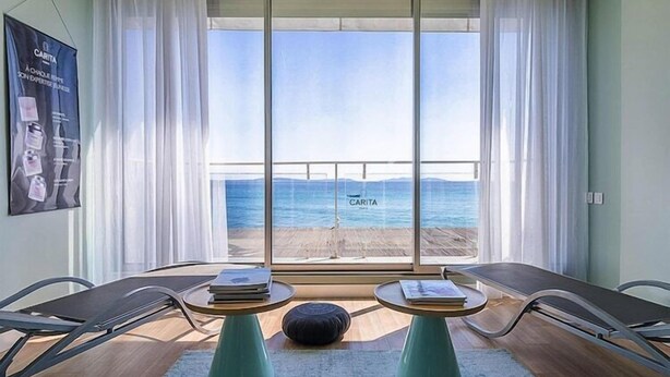 Une pièce avec une grande fenêtre donnant sur une plage, deux tables avec des livres et une affiche accrochée au mur.