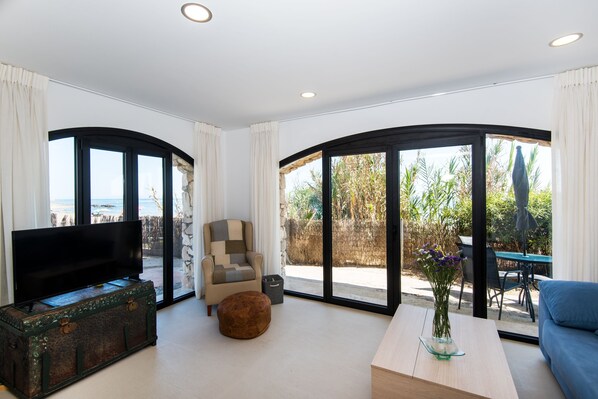 A modern living room with a flat-screen TV, a sofa, a coffee table, and a view of the outdoors through large glass doors.