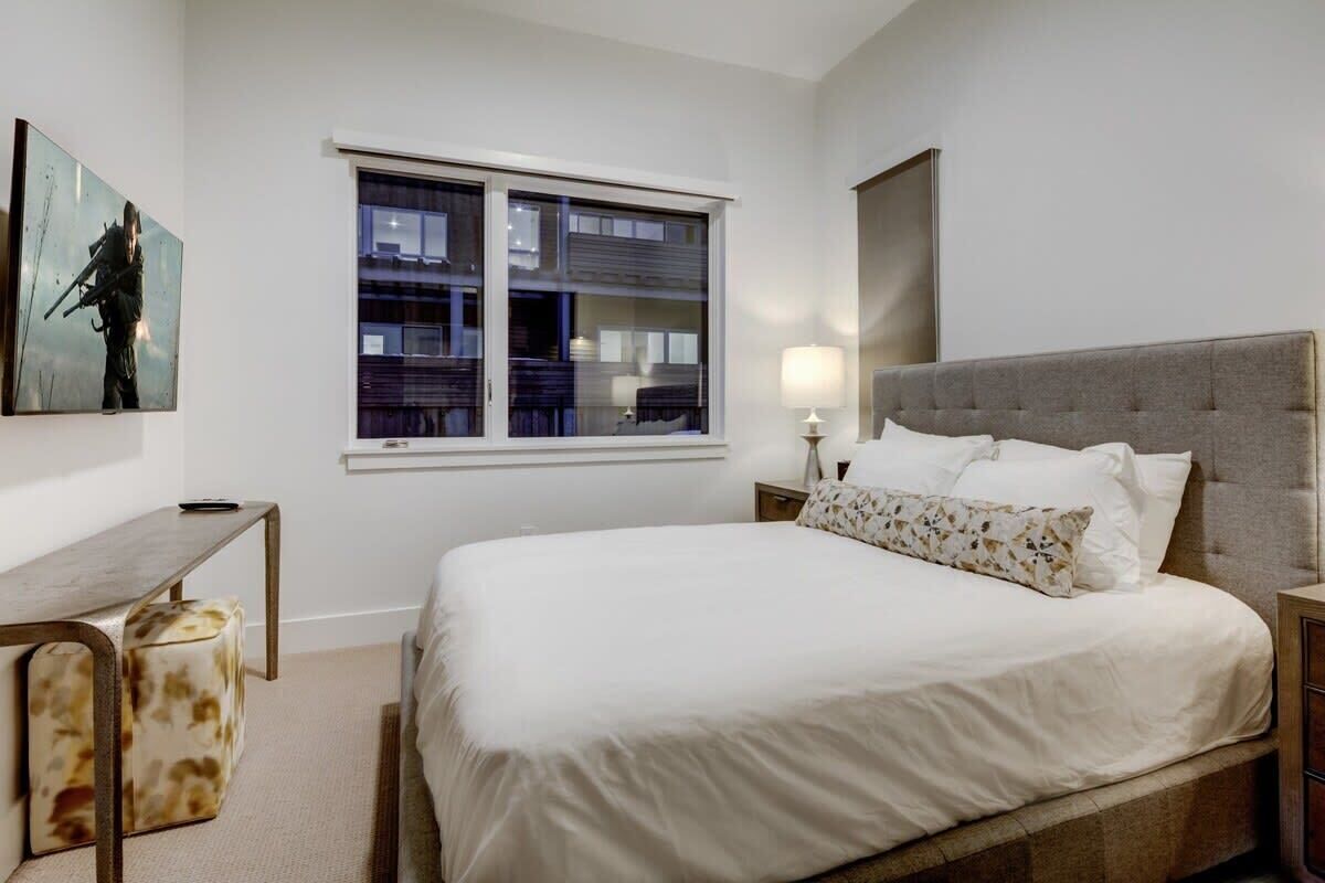 A bedroom with a large bed, a nightstand, a desk, a chair, and a window with blinds.