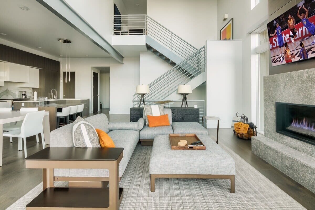 Ein modernes Wohnzimmer mit einem grauen Ecksofa, einem Couchtisch und einem Kamin. Eine Treppe führt zu einer oberen Ebene.
