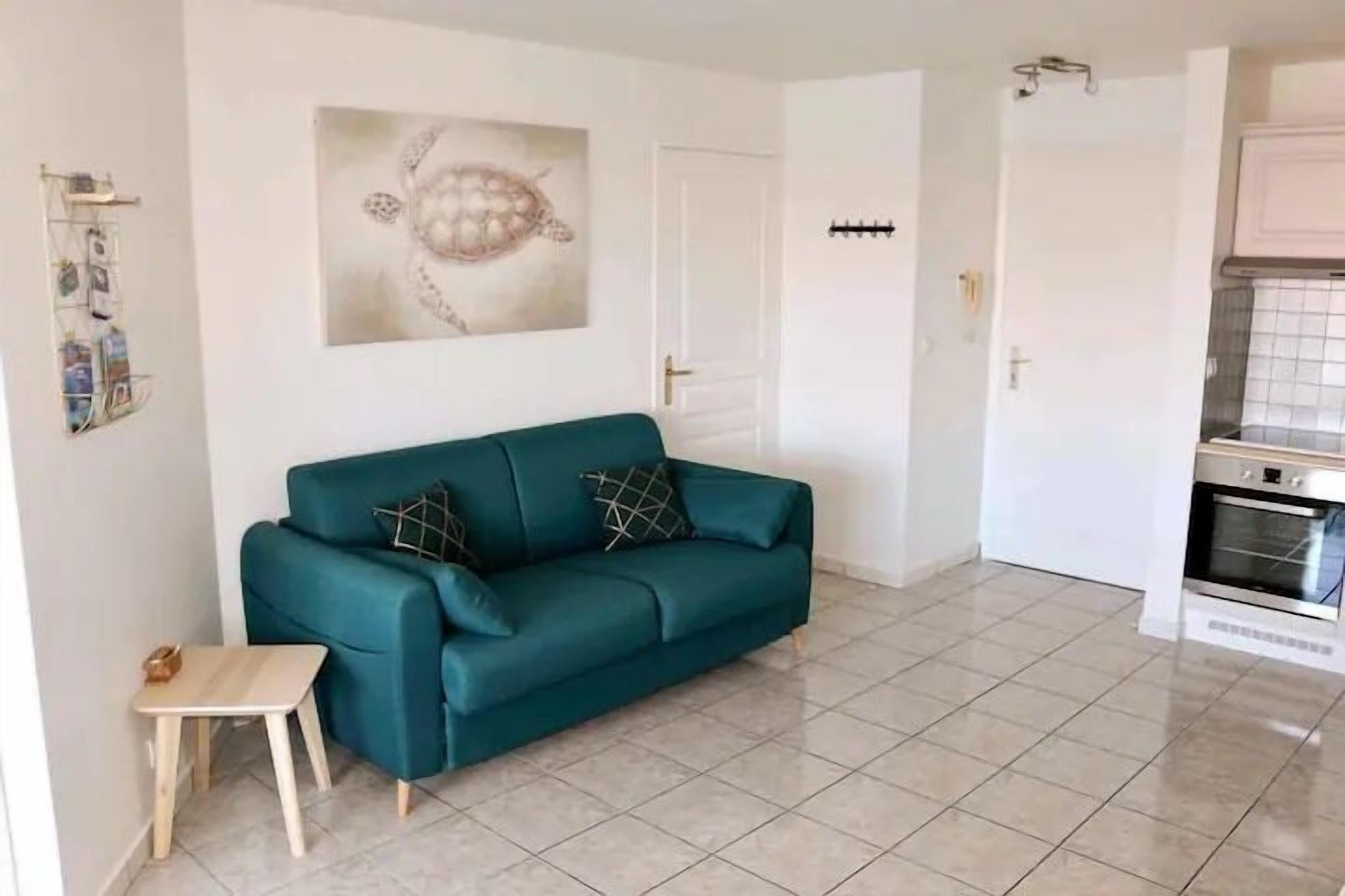 A teal sofa with patterned cushions, a small wooden table, a wall-mounted shelf with photos, and a kitchen area with a tiled backsplash and oven.