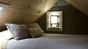A bedroom with a sloped ceiling, a window with a white lamp, and a bed with a quilted blanket and pillows. - Woodhaven Hideaway on Woods Pond.. (Bridgton)