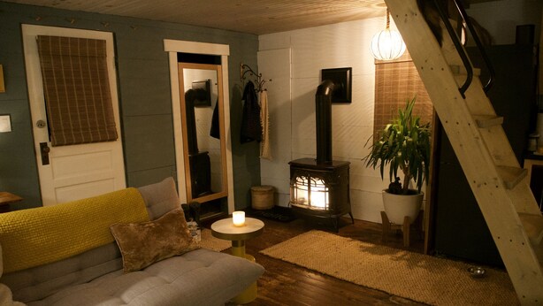 A cozy living room with a sofa, a small table with a candle, a wooden floor, a staircase, and a wood-burning stove. - Woodhaven Hideaway on Woods Pond.. (Bridgton)