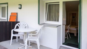 Un petit espace extérieur aménagé avec une table blanche et deux chaises en plastique blanches, une fenêtre avec un rideau et une porte donnant sur une pièce intérieure équipée d’un four à micro-ondes et d’une table en bois.