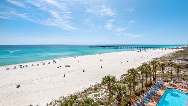 A beach resort with a long pier extending into the ocean, a sandy beach, and a pool area with lounge chairs.