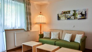 Un salon avec un canapé vert, trois tables en bois, un radiateur, une lampe et trois tableaux accrochés au mur.