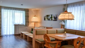 Un coin repas avec une table et des chaises en bois, un banc servant de siège, et un radiateur placé sous une fenêtre ornée de rideaux légers.