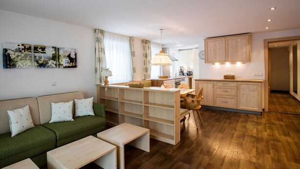 Un salon moderne avec un canapé vert, une table basse en bois et un coin repas comprenant une table et des chaises. On y trouve également une cuisine équipée de placards et d’un luminaire suspendu.