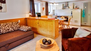 Ein gemütliches Wohnzimmer mit einer Couch, einem Holztisch mit Dekoration, einem roten Sessel und einer Küchenzeile mit Schränken und einem Kühlschrank.