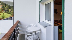 Un balcon avec une balustrade en bois, une table blanche et deux chaises blanches, une fenêtre et une vue sur un paysage verdoyant, avec des arbres et des fleurs rouges.