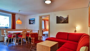 A living room with a red sofa, wooden dining table, and chairs. There are two paintings on the wall and a window with curtains.