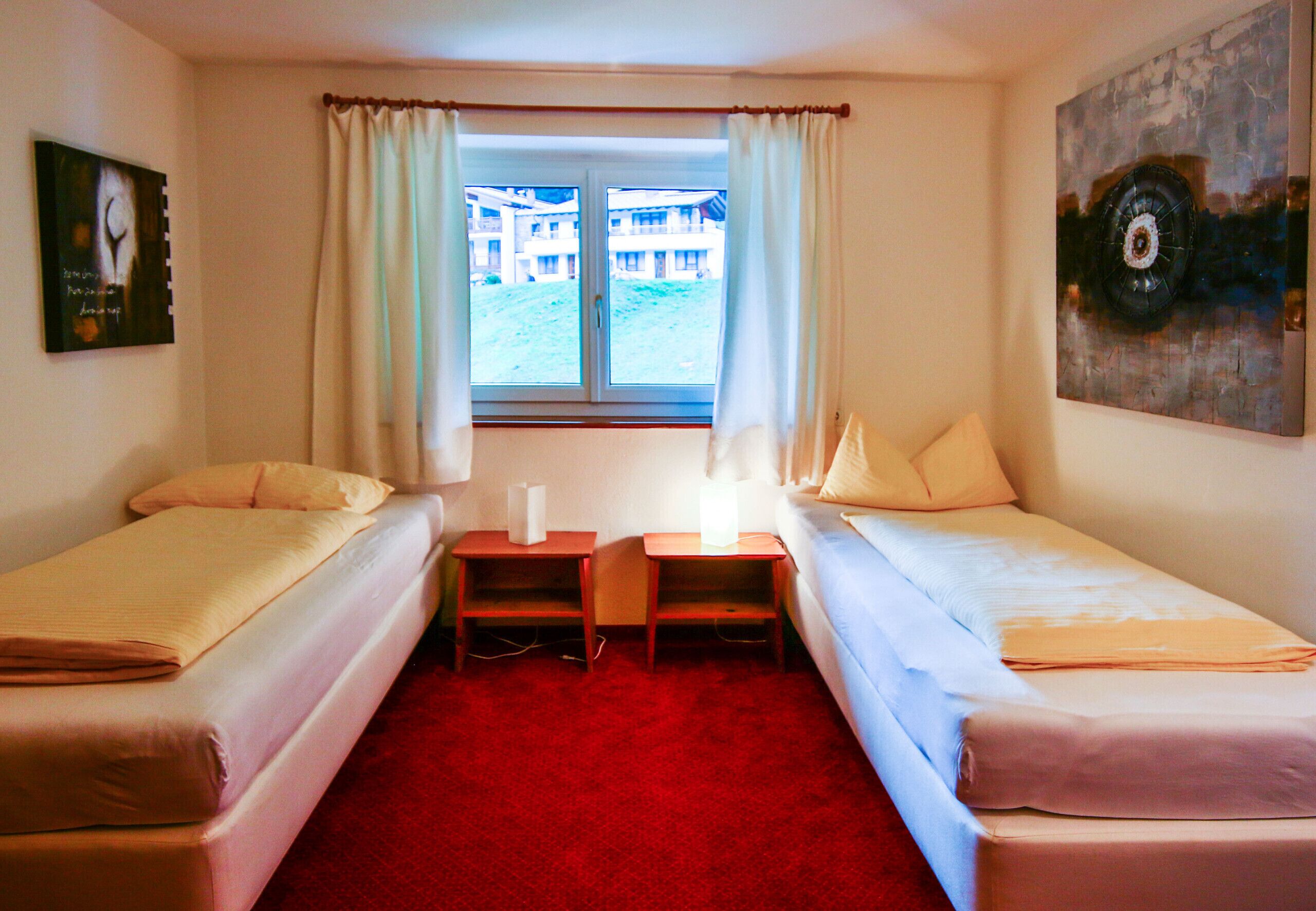 Een kamer met twee eenpersoonsbedden, een tafeltje, een lamp, een schilderij en een raam met gordijnen.
