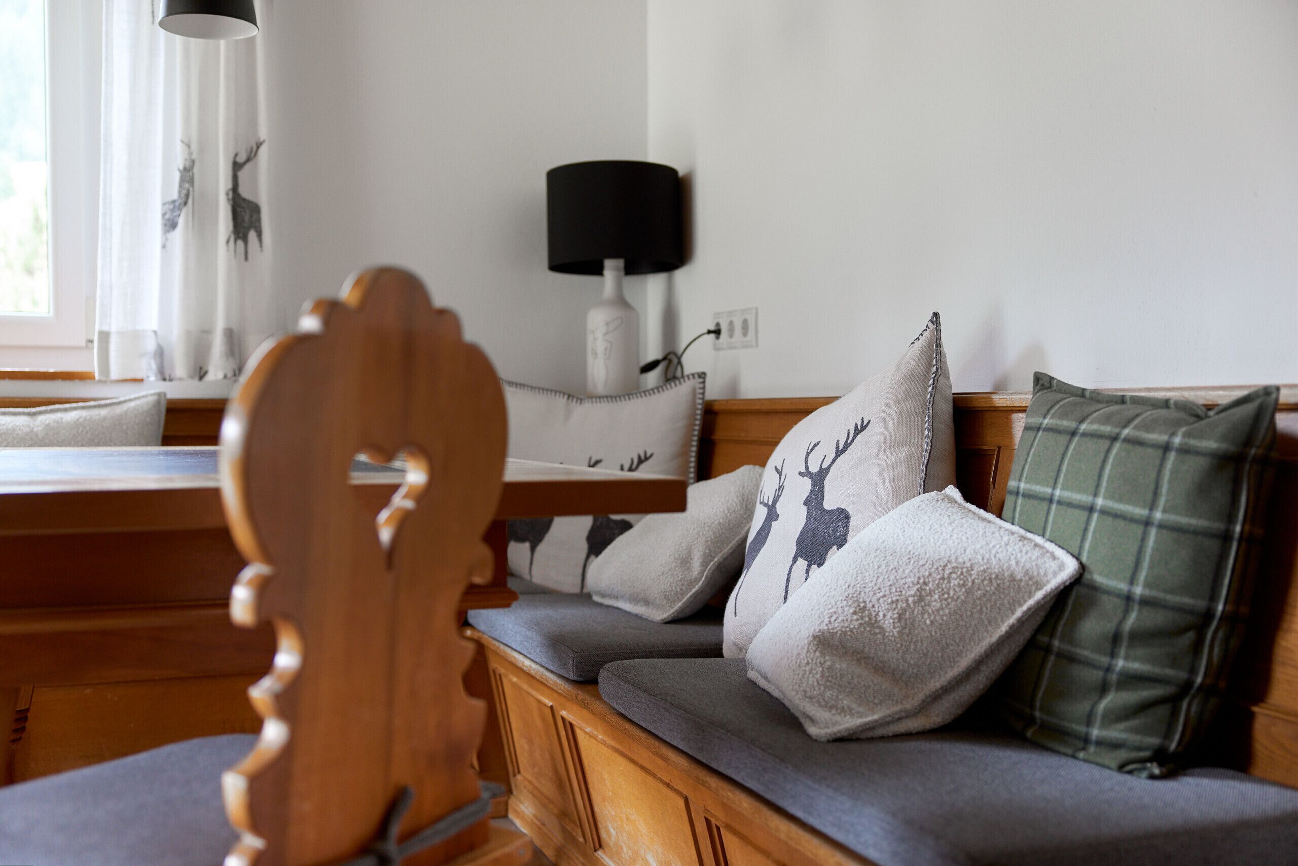 Un banc en bois avec des coussins gris et des oreillers décoratifs, une lampe blanche et une fenêtre avec des rideaux.