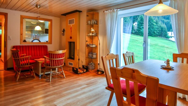A cozy room with a wooden ceiling, a dining table with chairs, a red sofa, and a large window offering a view of the outdoors.