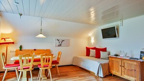 A room with a wooden dining table and chairs, a bed with red and white bedding, a wooden cabinet, and a wall-mounted television.