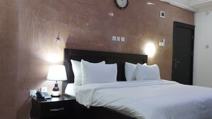 A hotel room with a bed, a nightstand with a lamp, a clock, and a door. - Peace Media Hotels Gwarimpa (Abuja)
