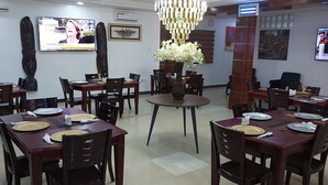 A dining area with multiple tables and chairs, a central round table with a vase of flowers, and two televisions mounted on the walls displaying different programs. - Peace Media Hotels Gwarimpa (Abuja)